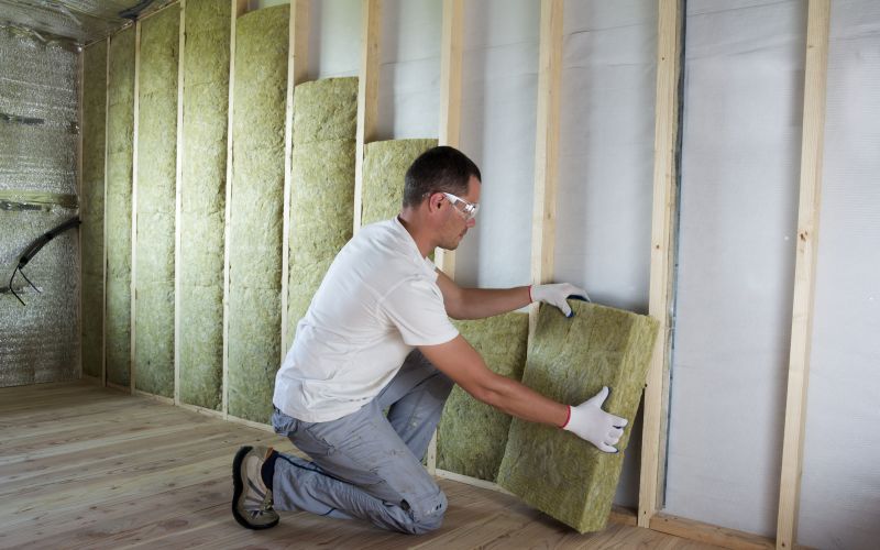 Worker in protective goggles and respirator insulating rock wool insulation in wooden frame for future house walls